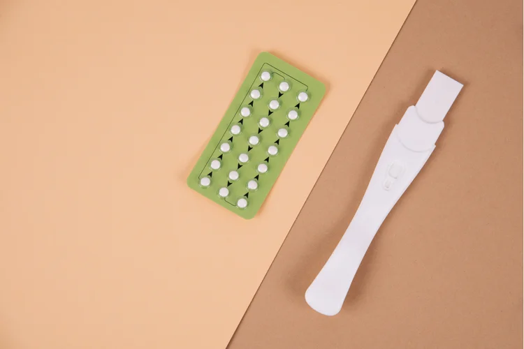 Green pack of oral contraceptive pills next to pregnancy test on beige and brown background.