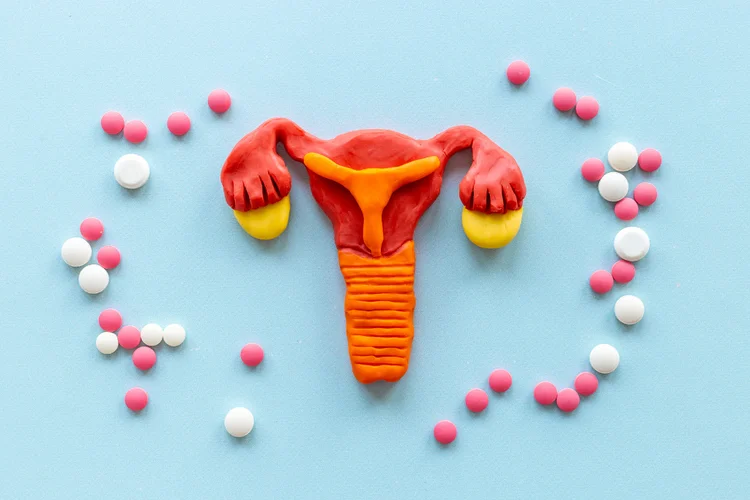 Uterus model made of clay surrounded by pink and white oral contraceptive pills on blue background.